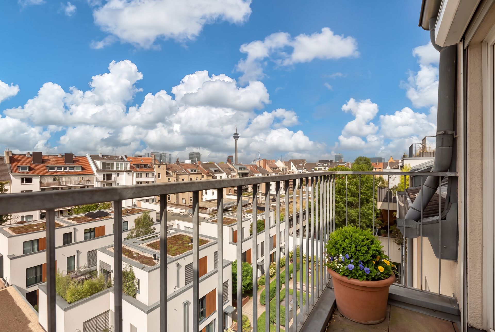 Blick von einem Balkon mit geländer und Blumentopf auf Wohnhäuser und blauen Himmel.