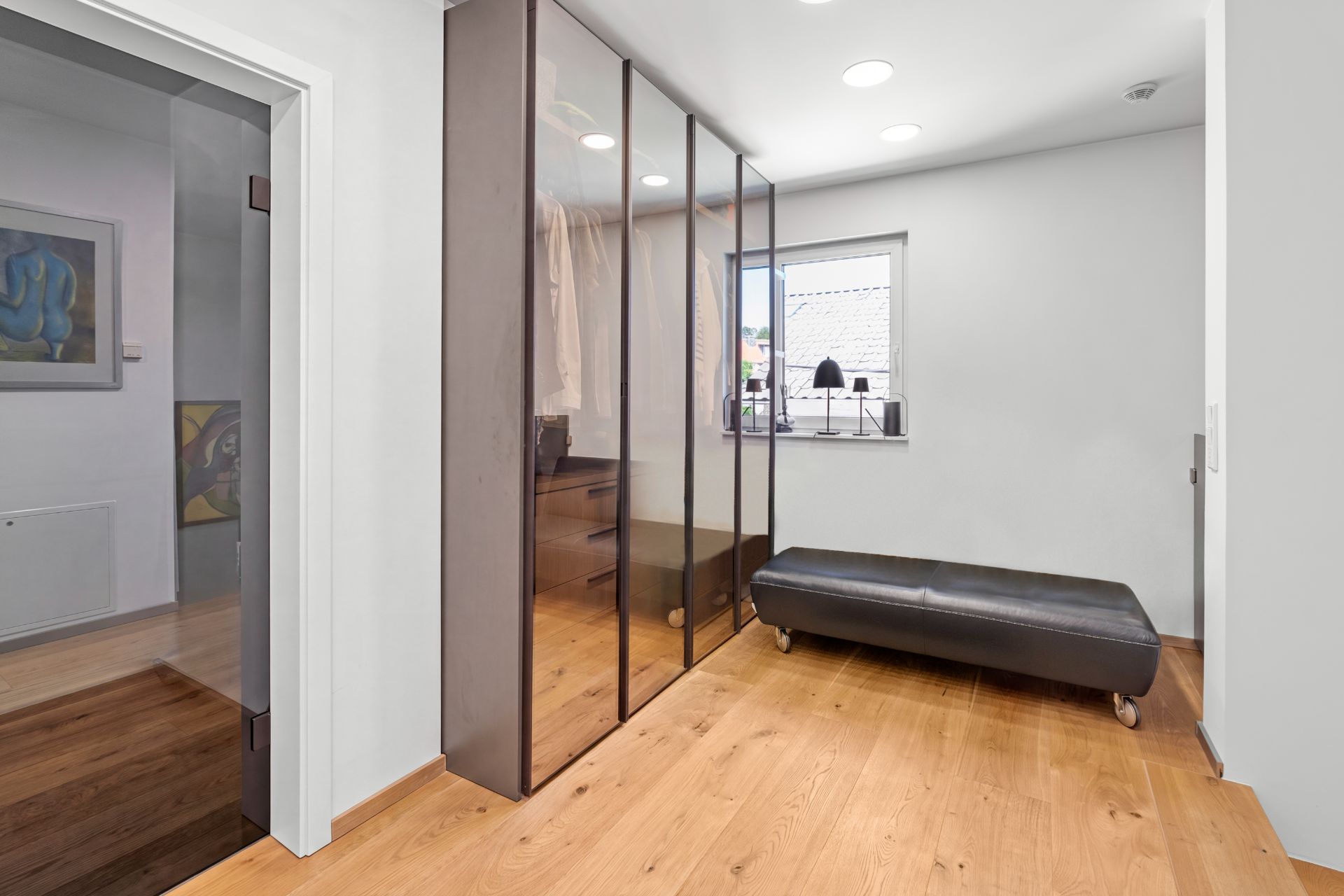 Moderner Ankleideraum mit Spiegelschrank, Holzfußboden und Sitzbank vor einem Fenster.