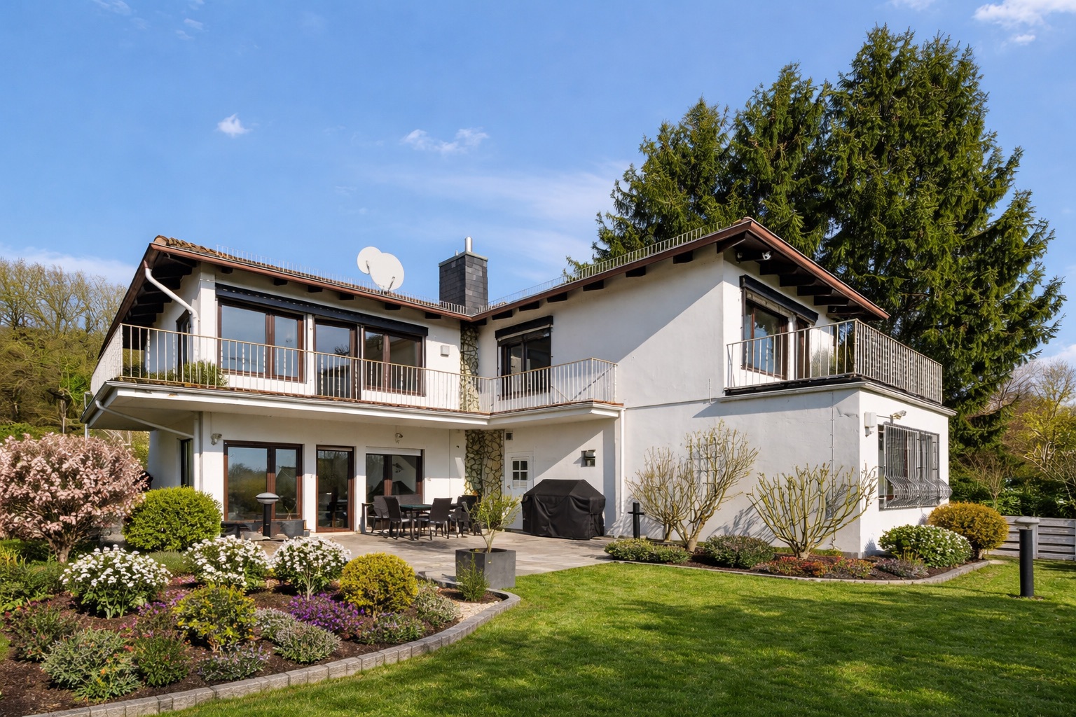 Modernes zweistöckiges Wohnhaus mit Balkon, Terrasse und gepflegtem Garten bei Sonnenschein.