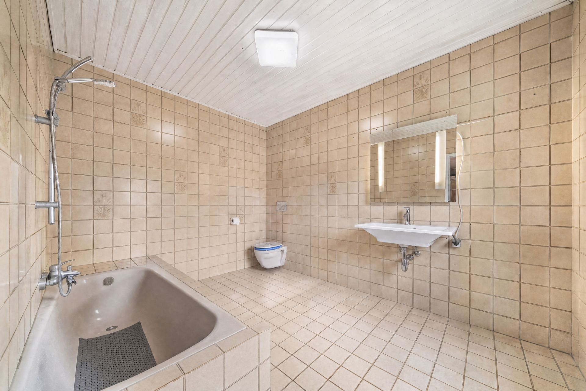 Großes Bad mit hellen Fliesen, Badewanne, Wand-WC und Waschbecken mit Spiegel.