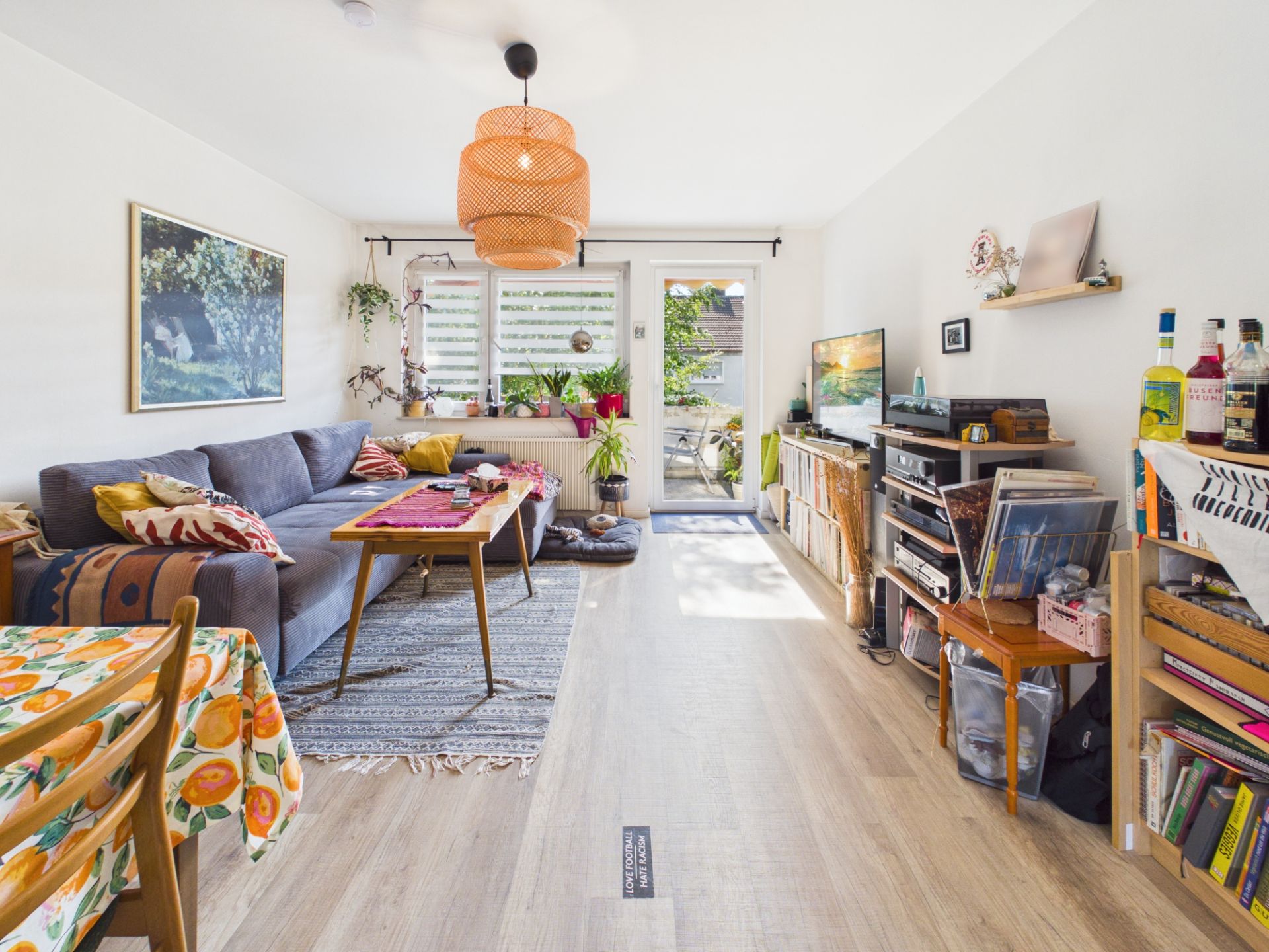 Helles Wohnzimmer mit grauem Sofa, Esstisch, Regalen, Pflanzen und Zugang zu einem Balkon.