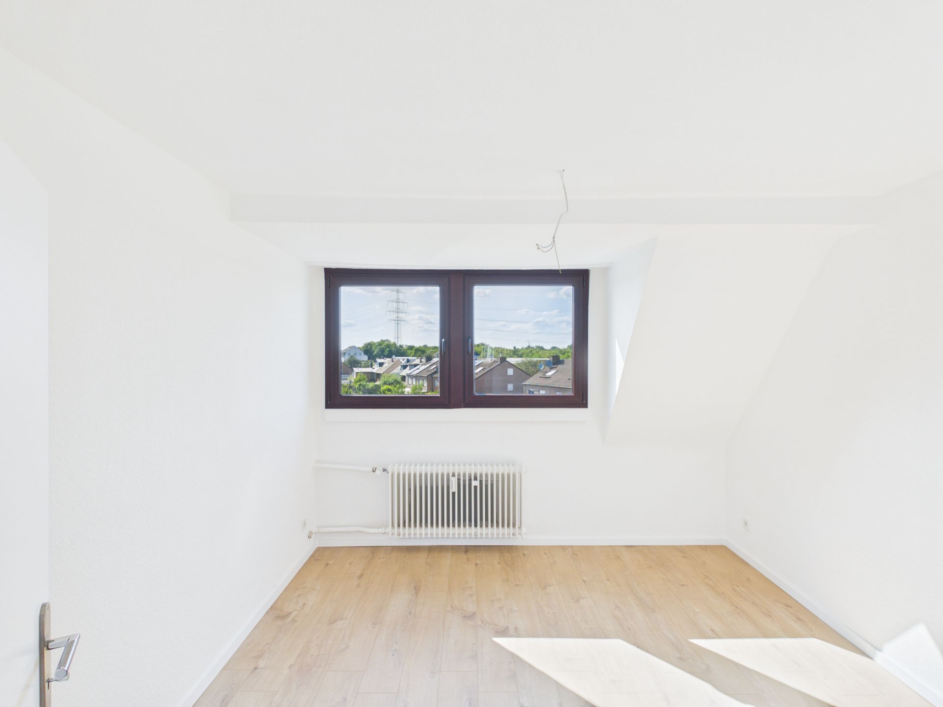 Helles, leeres Zimmer mit Holzfußboden, großem Fenster und Heizkörper unter dem Fenster.