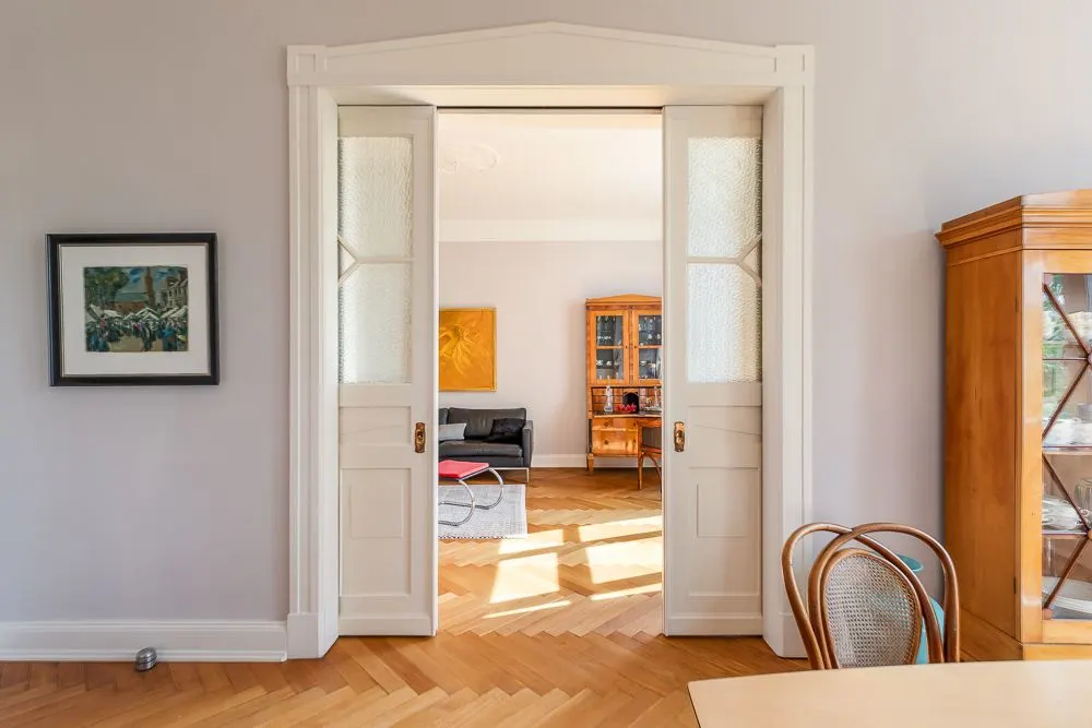 Blick durch geöffnete Flügeltüren auf helles Wohnzimmer mit Holzschränken und Sofa.
