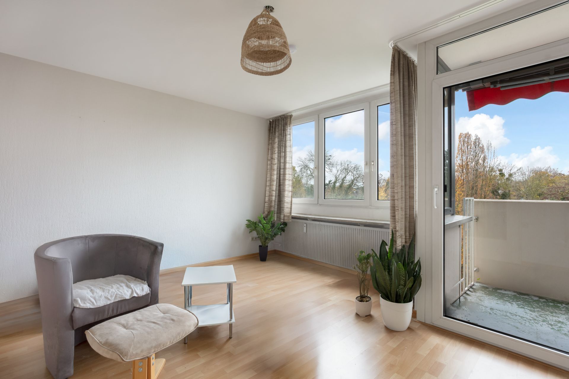 Helles Wohnzimmer mit Sessel, Fußhocker, kleinem Tisch, Pflanzen und Zugang zum Balkon.