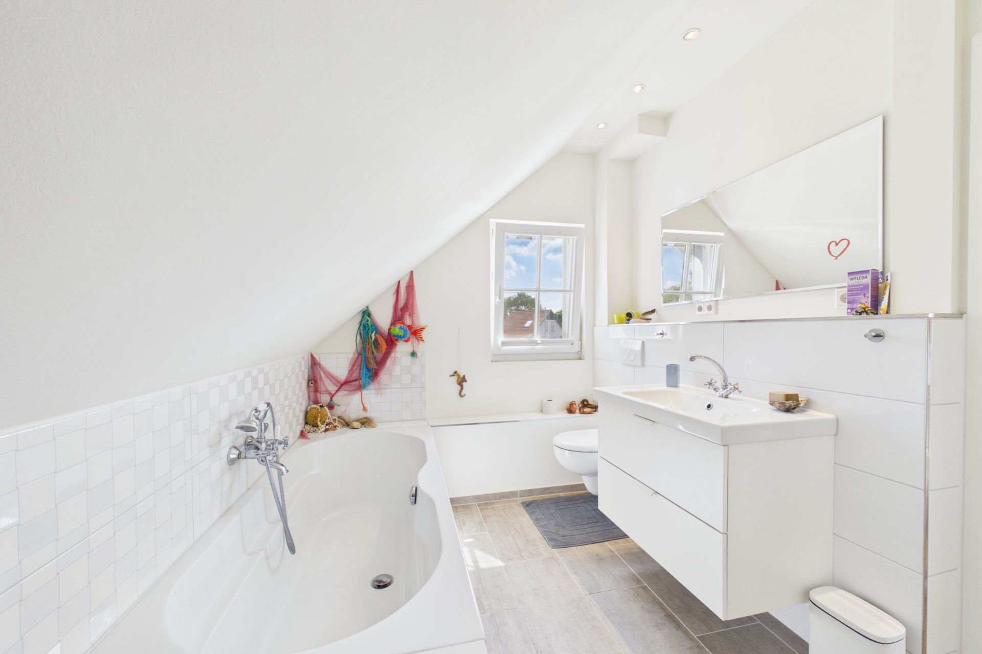 Helles Badezimmer mit Badewanne, Waschbecken, Spiegel und Fenster unter einer Dachschräge.
