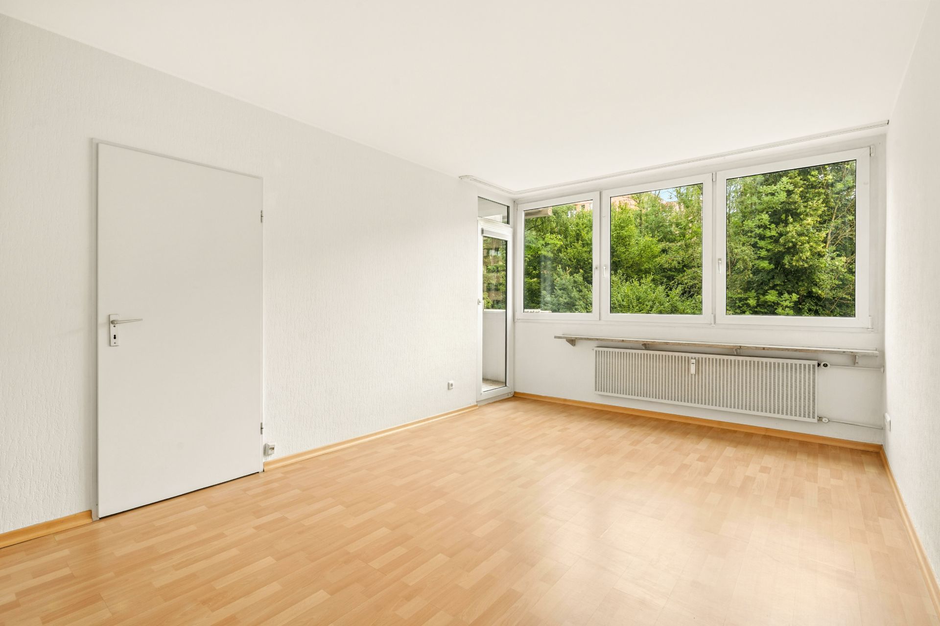 Leerer Raum mit hellem Holzboden, weißen Wänden und großen Fenstern mit Blick ins Grüne.
