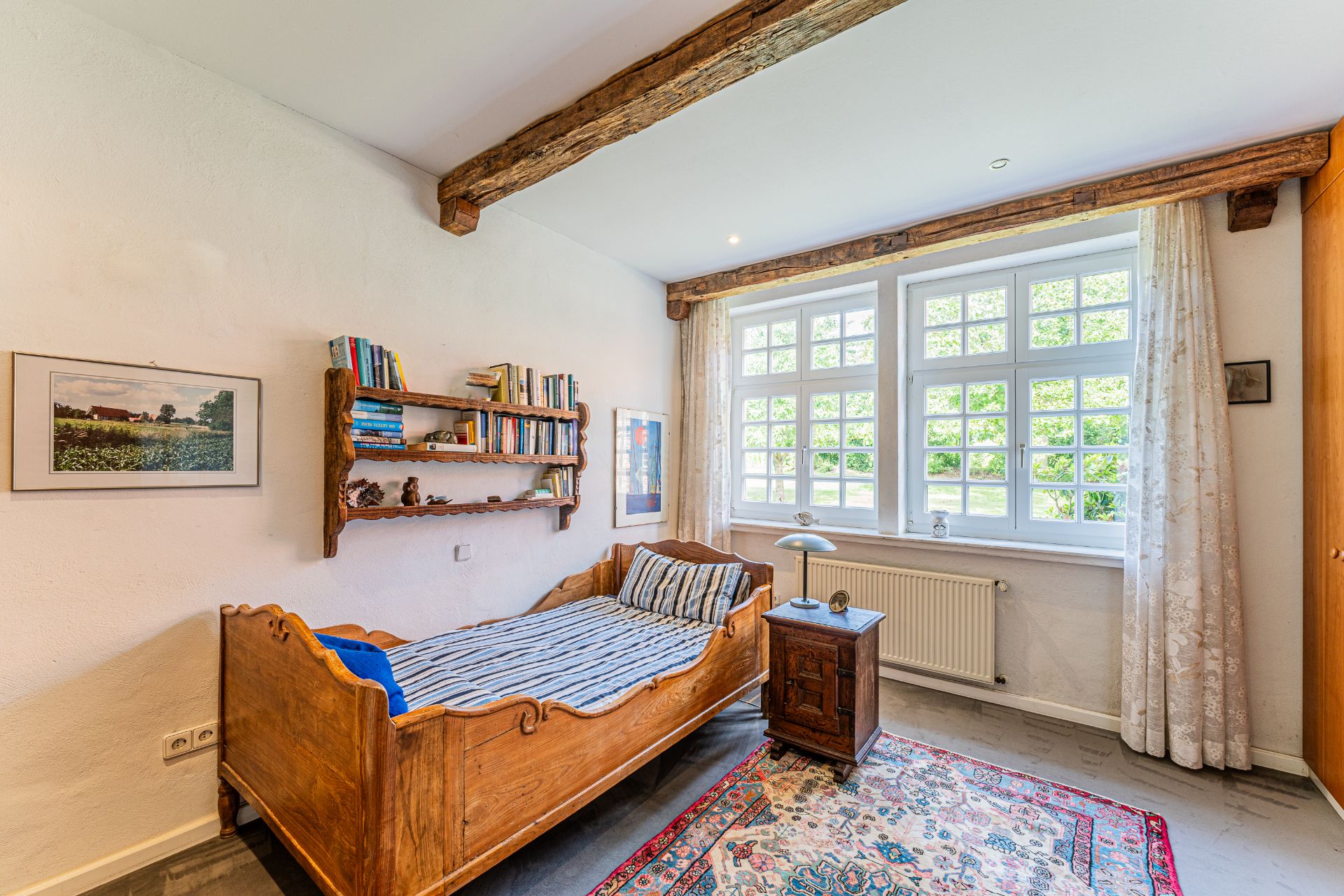 Helles Schlafzimmer mit Holzbett, Bücherregal, Teppich und großem Fenster mit Vorhängen.