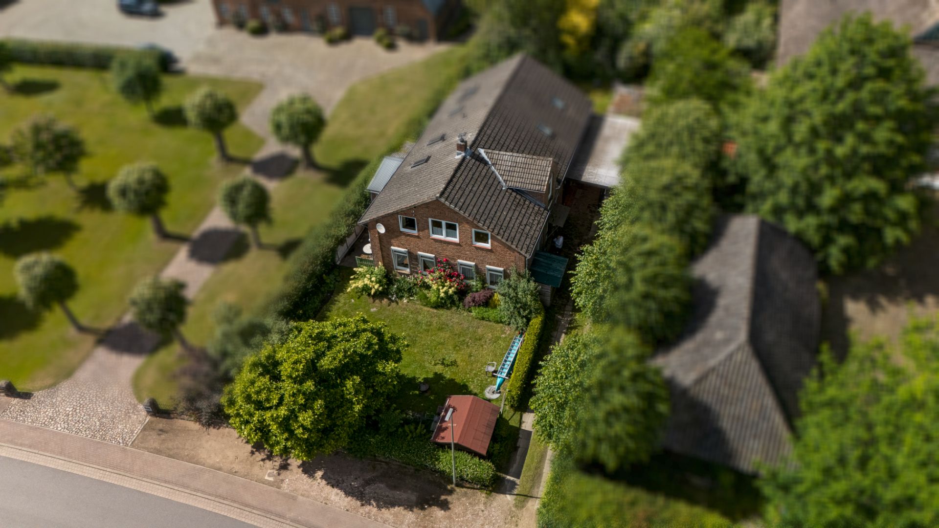 Luftaufnahme eines Einfamilienhauses mit Garten und Bäumen an einer Straße.