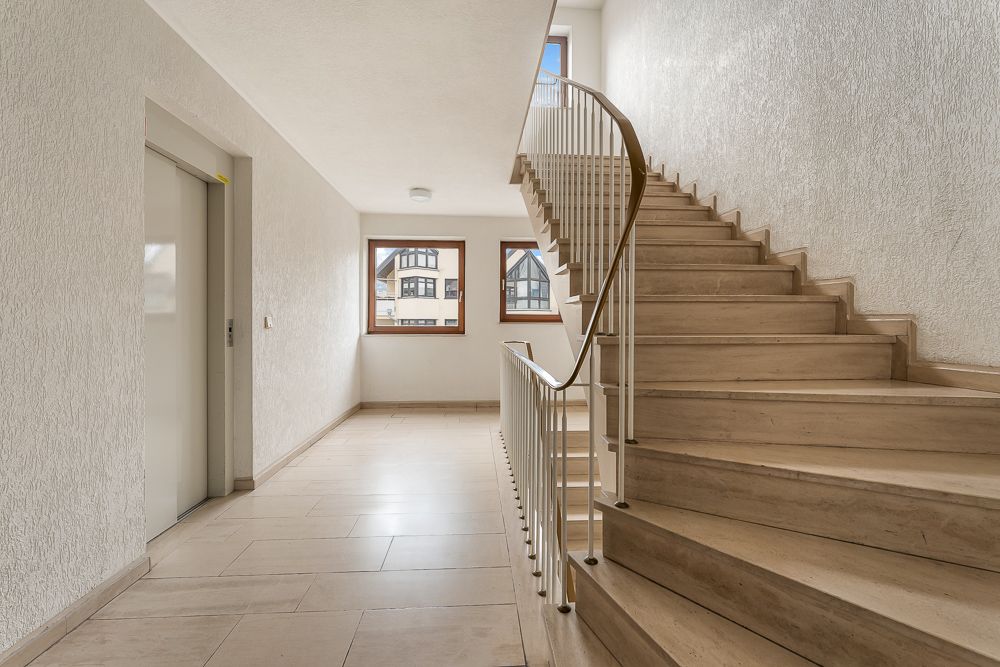 Heller Hausflur mit Aufzug, Treppenhaus und zwei Fenstern mit Blick nach draußen.