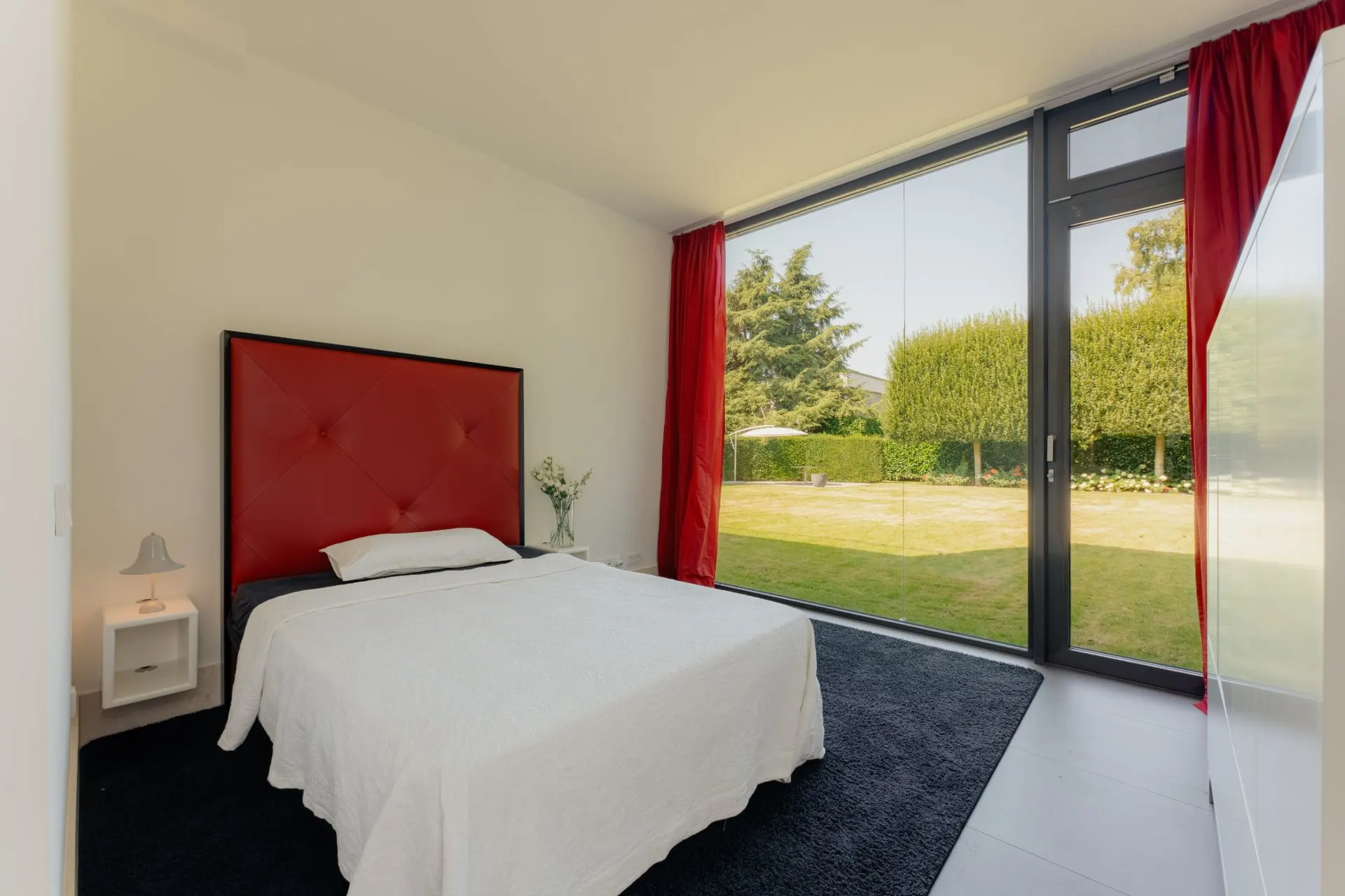 Schlafzimmer mit großem Fenster, rotem Kopfteil, rotem Vorhang und Blick auf Garten.