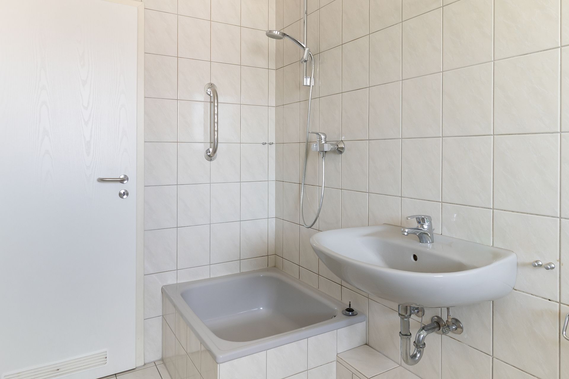 Badezimmer mit weißem Waschbecken, Dusche, Haltegriff und gefliestem Boden.
