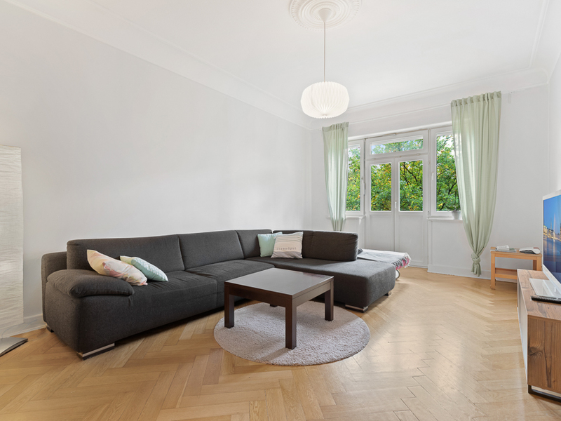Helles Wohnzimmer mit großer grauer Eckcouch, Couchtisch und Fenstern mit grünen Vorhängen.