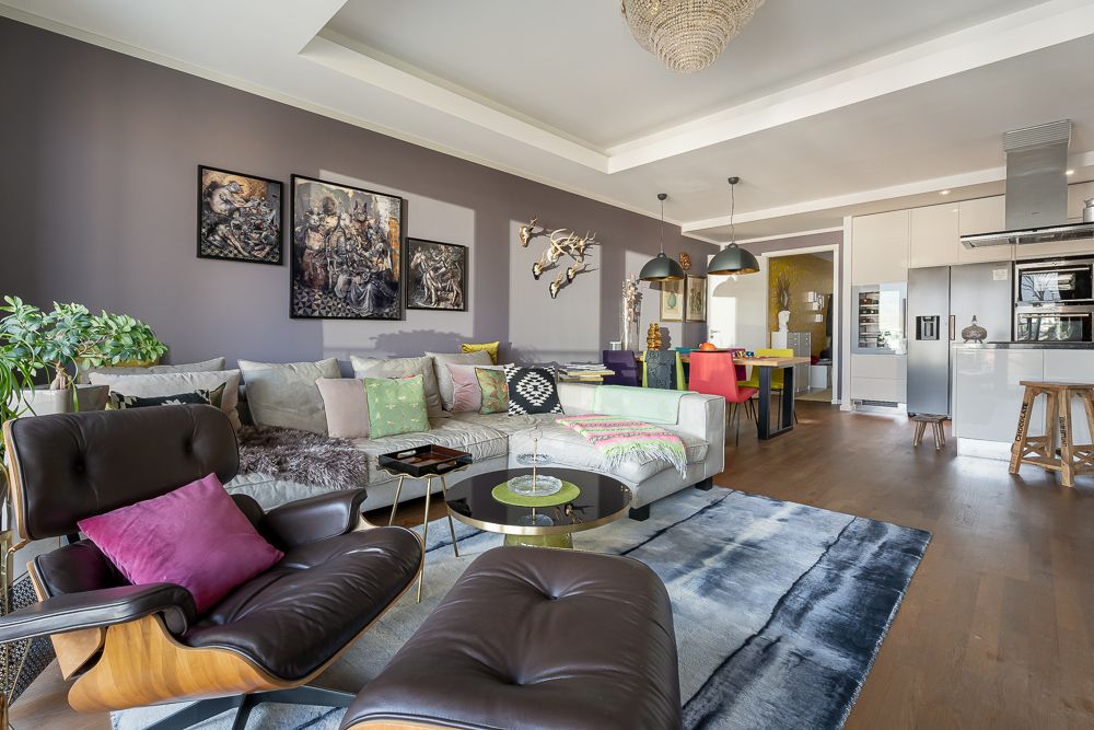 Modernes Wohnzimmer mit Sofa, Sesseln, Teppich, Pflanzen und offener Küche im Hintergrund.