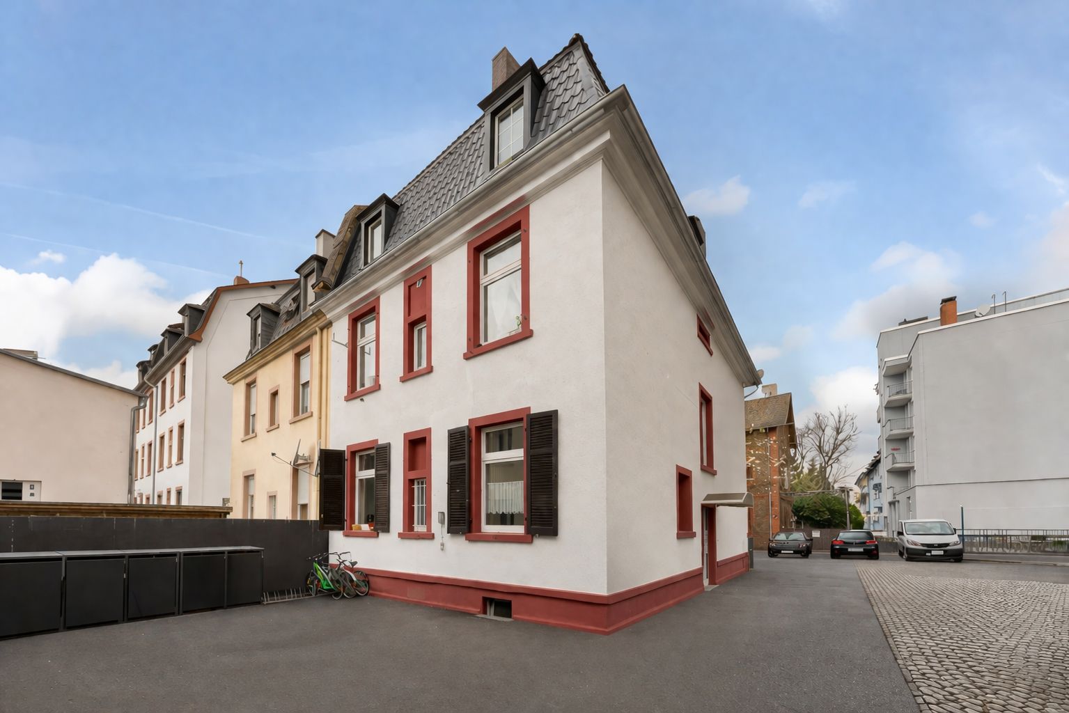 Mehrstöckiges Wohnhaus mit weißen Wänden und roten Fensterrahmen, davor Asphaltfläche und parkende Autos.