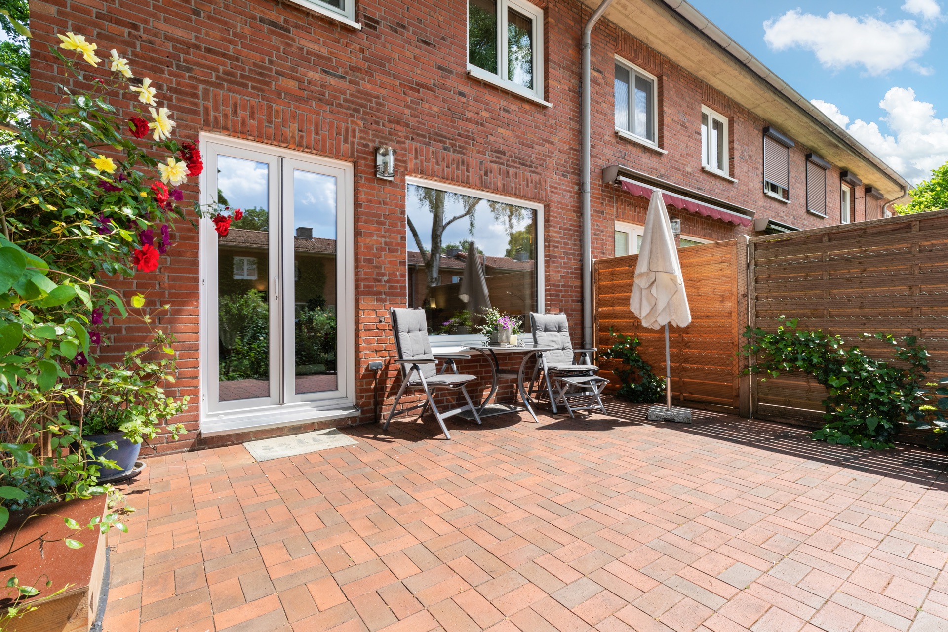 Gepflasterte Terrasse mit Tisch, zwei Stühlen, Sonnenschirm und Blumen vor Backsteinhaus.