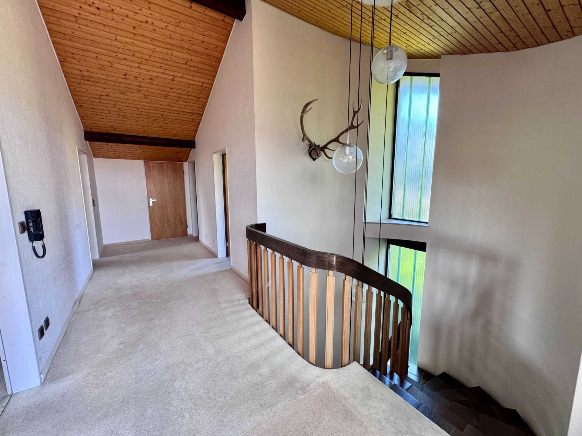 Heller Flur mit Teppichboden, Holzdecke, Treppe, Geweih an der Wand und großen Fenstern.