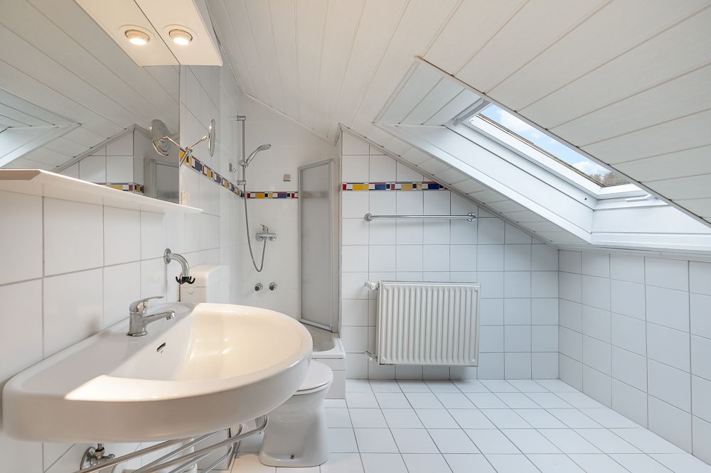 Helles Badezimmer mit Dachschräge, Waschbecken, Toilette, Dusche und großem Fenster.