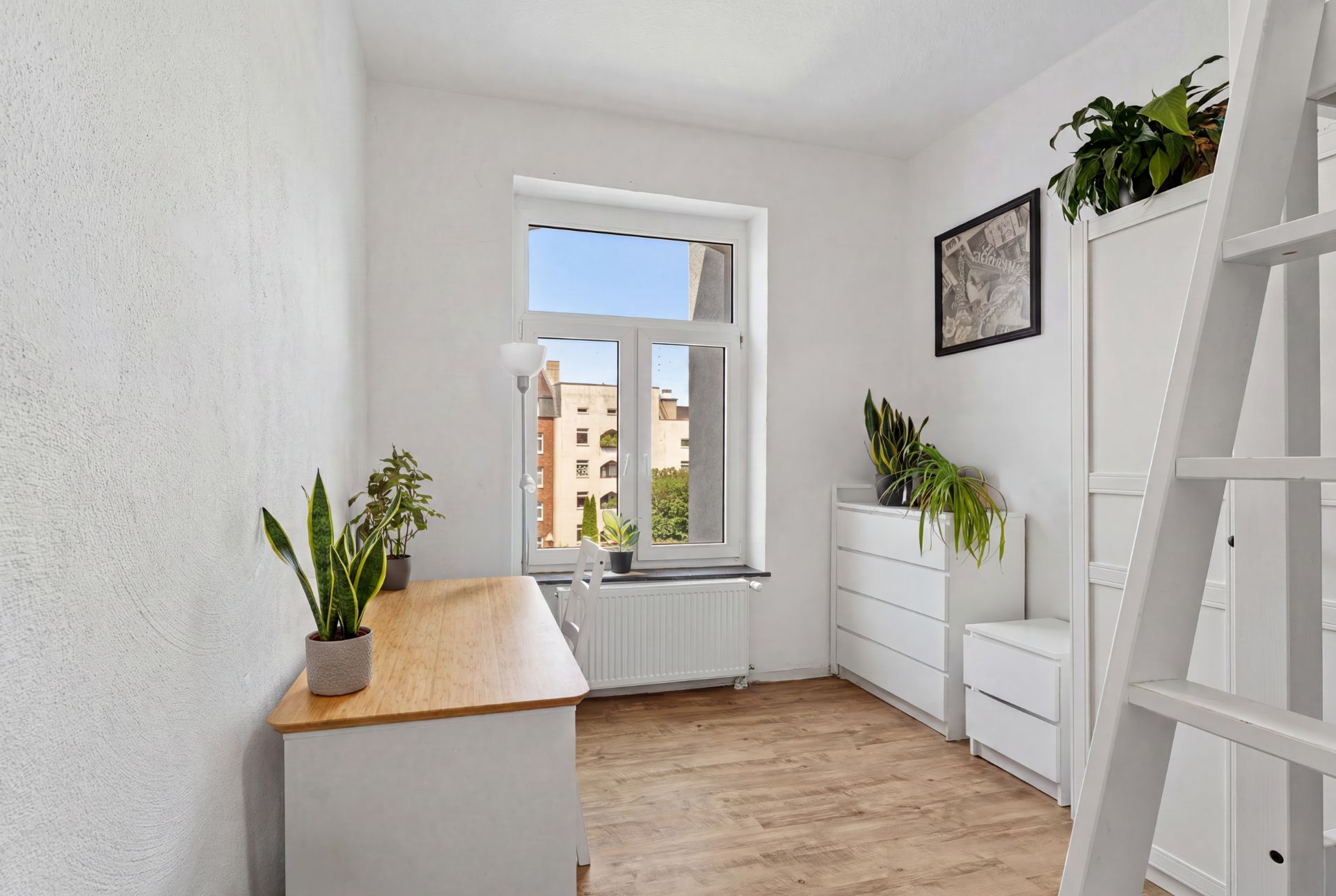 Helles Zimmer mit Pflanzen, Schreibtisch, Kommoden und großem Fenster mit Blick nach draußen.