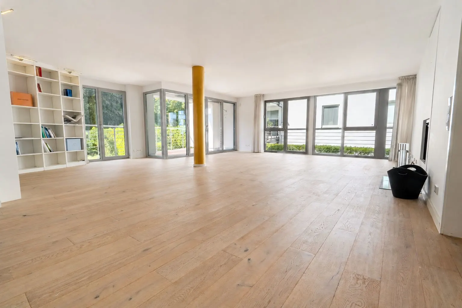 Heller, leerer Wohnraum mit Holzfußboden, großen Fenstern und Regalen an der Wand.
