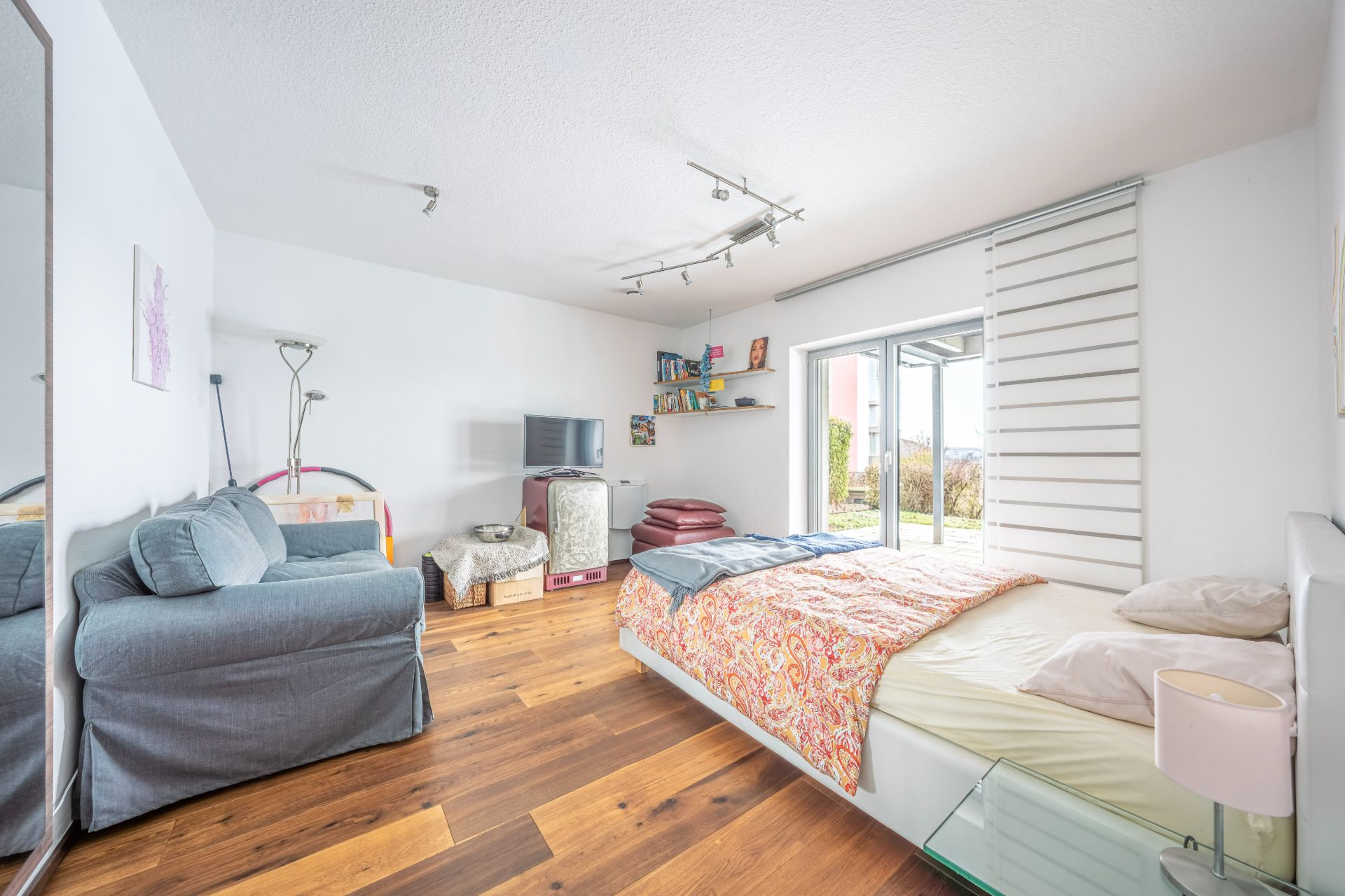 Helles Schlafzimmer mit großem Bett, grauer Couch, Fernseher und Zugang zur Terrasse.