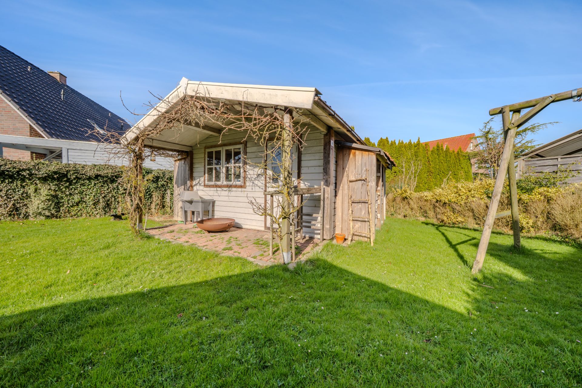 Kleines Holzhaus mit überdachter Terrasse in einem Garten mit Rasen und Schaukel.