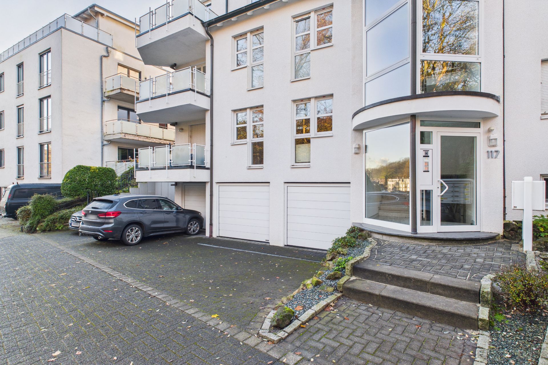 Eingang eines modernen Wohnhauses mit Garagen, Treppe und geparkten Autos davor.