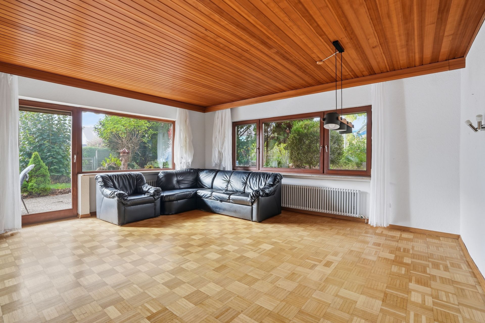 Helles Wohnzimmer mit Holzdecke, Parkettboden, großer schwarzer Ledercouch und bodentiefen Fenstern.