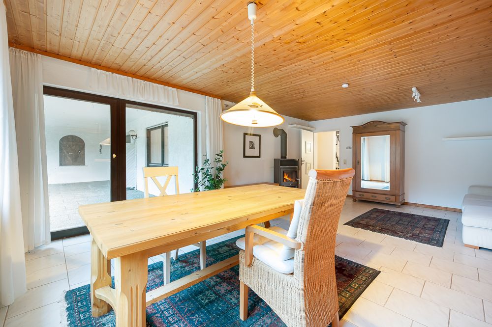 Helles Wohnzimmer mit Holzdecke, Esstisch, Kaminofen, Teppichen und großem Fenster.