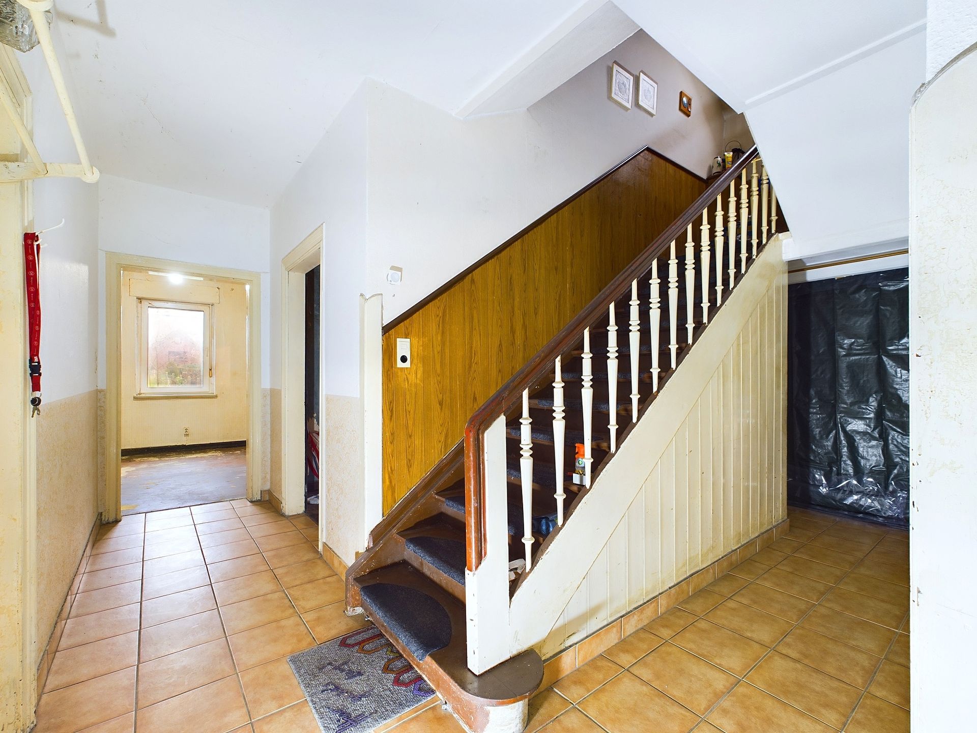 Treppenhaus mit braunen Fliesen, Holztreppe und Flur in einem Wohnhaus.