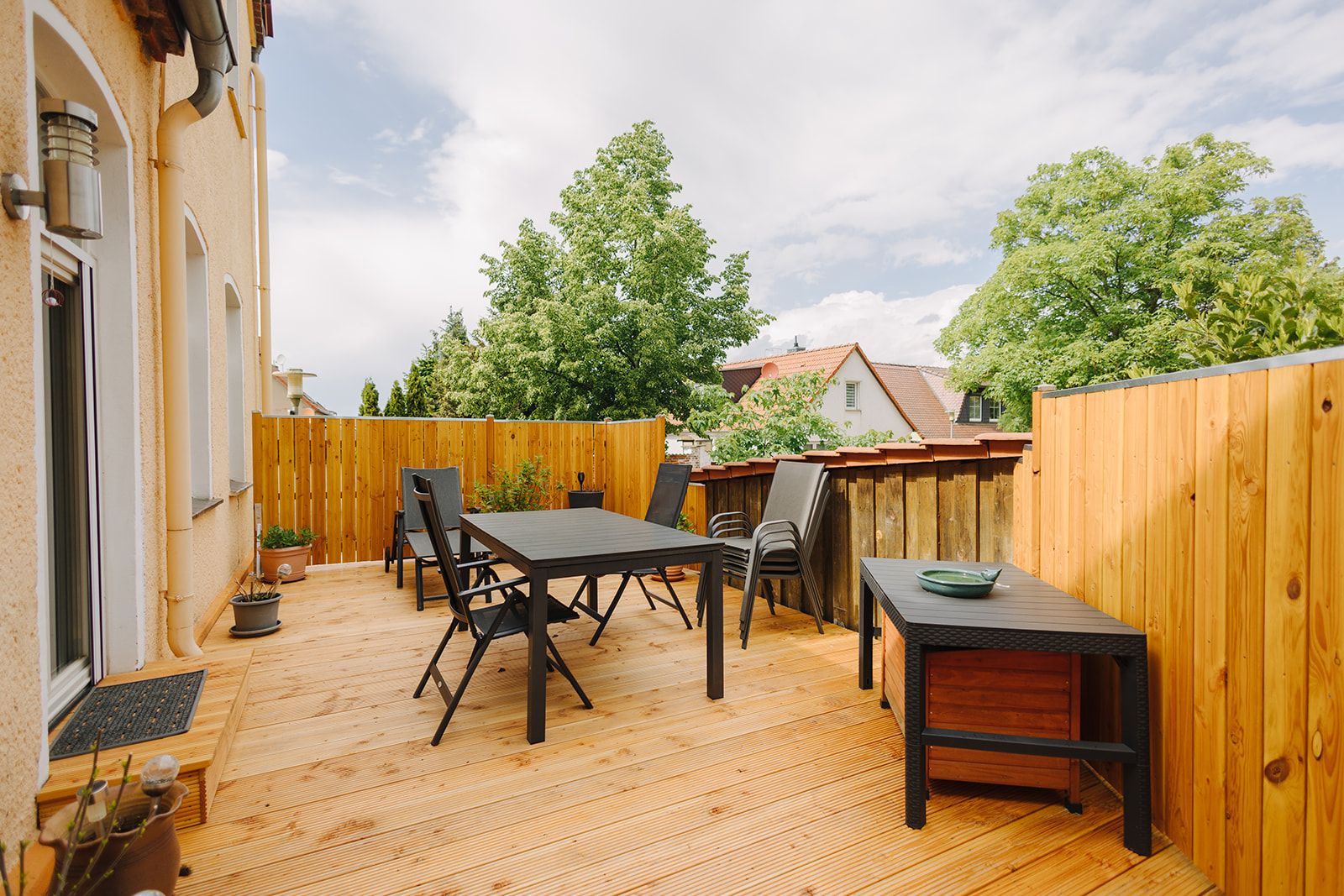 Holzterrasse mit Tisch, Stühlen und Zaun, daneben grüne Bäume und Nachbarhäuser.