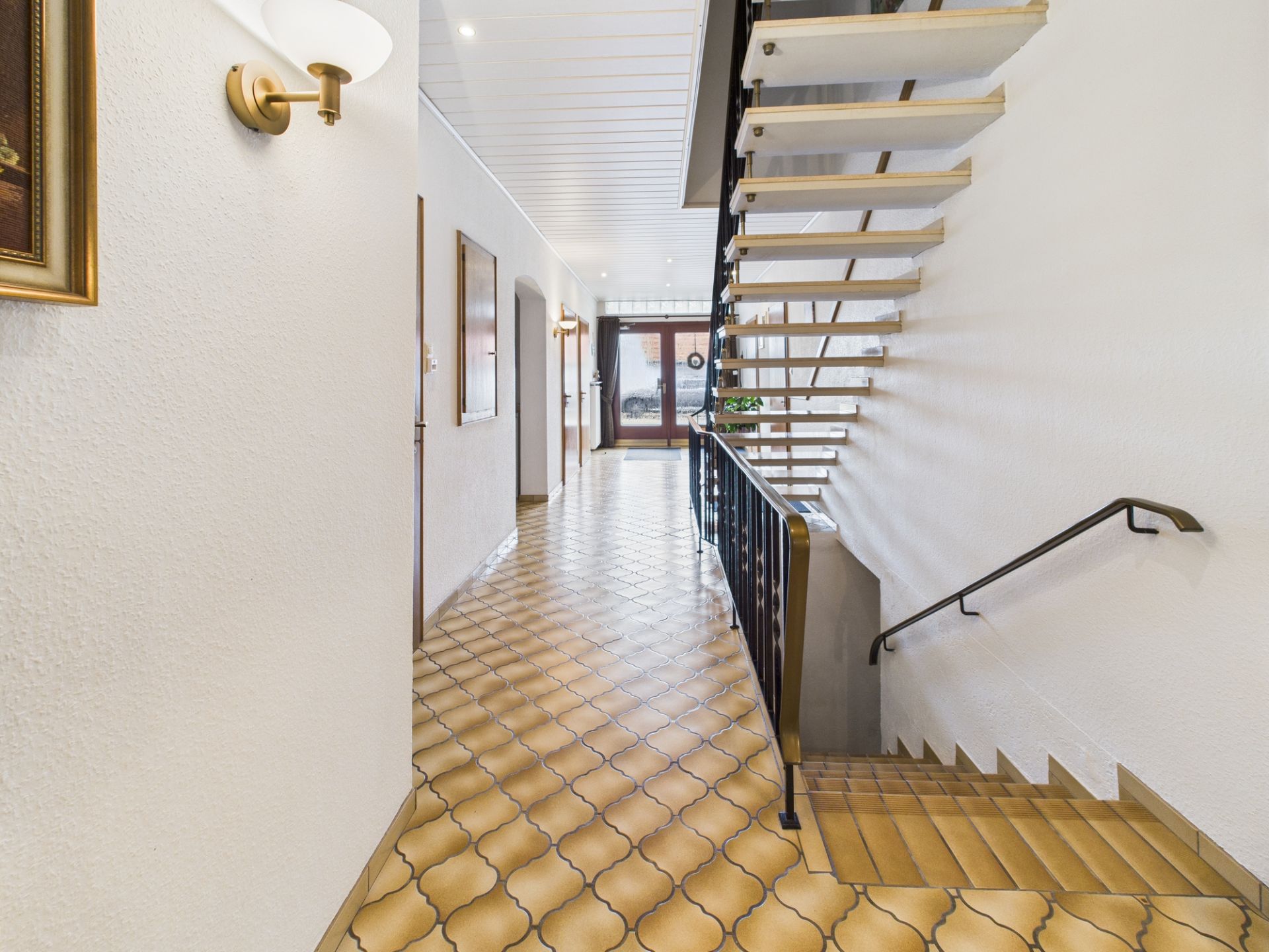 Heller Flur mit gemustertem Fliesenboden und einer offenen Treppe, Wandlampen und Bildern an den Wänden.