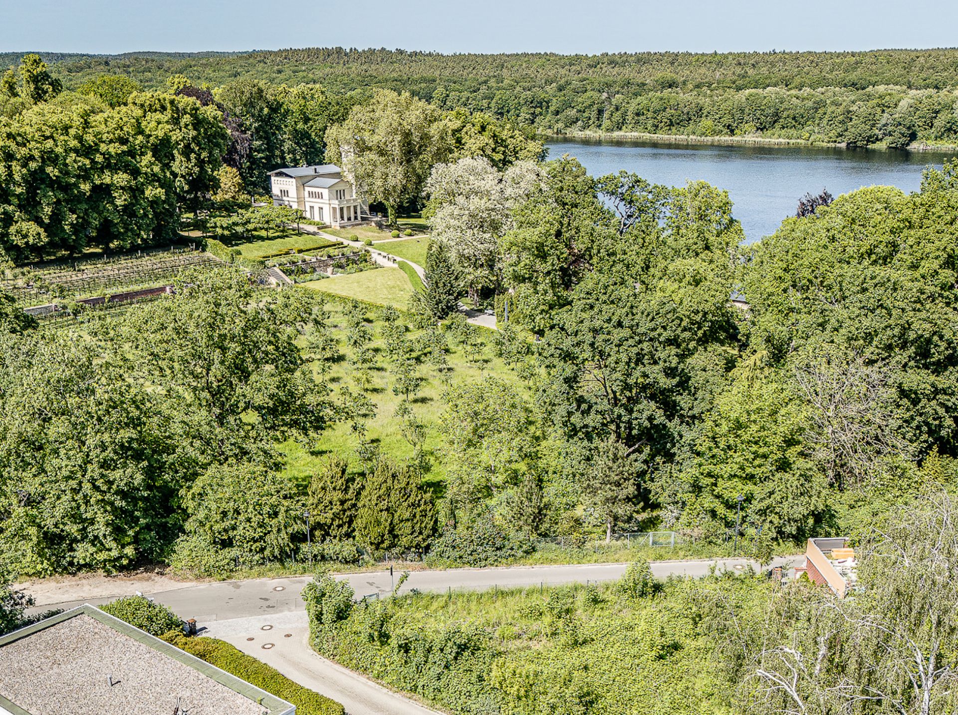 Luftaufnahme eines großen Gartens mit Bäumen, Rasen, Haus und angrenzendem See.