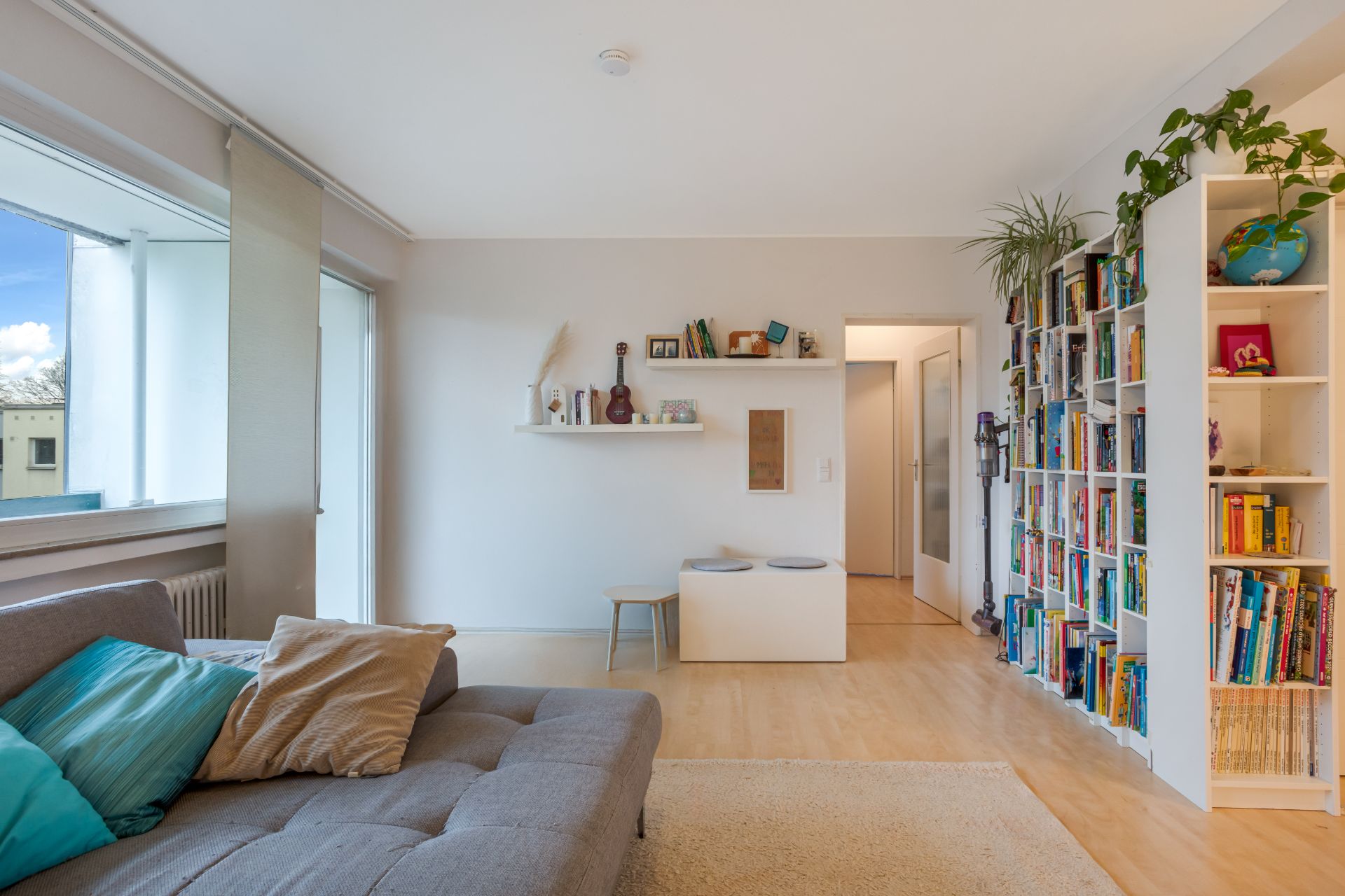 Helles Wohnzimmer mit grauem Sofa, Bücherregal, Fenster und Wandregalen mit Dekoration.