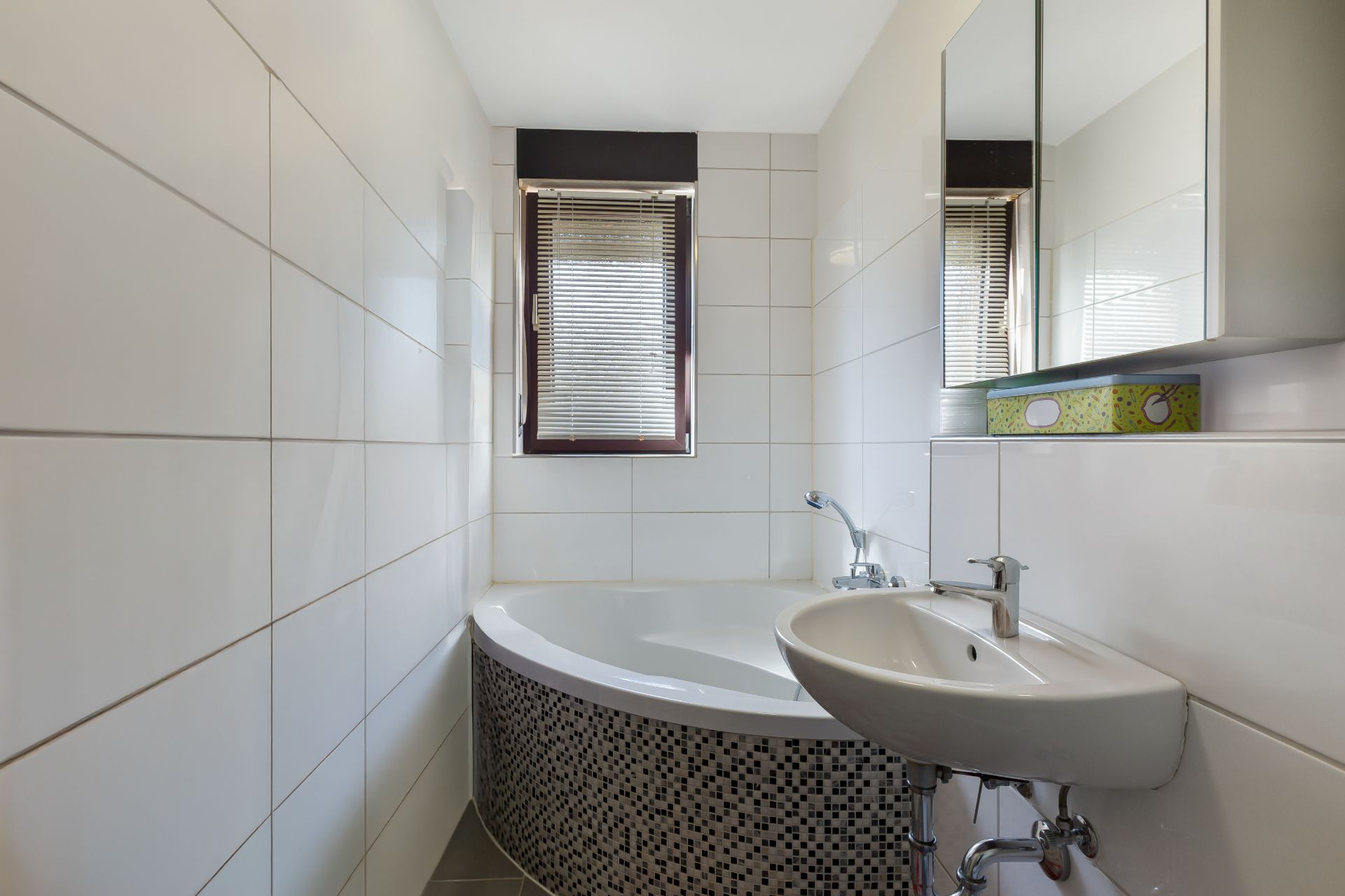 Kleines Badezimmer mit Eckbadewanne, Waschbecken und Fenster mit Jalousien.