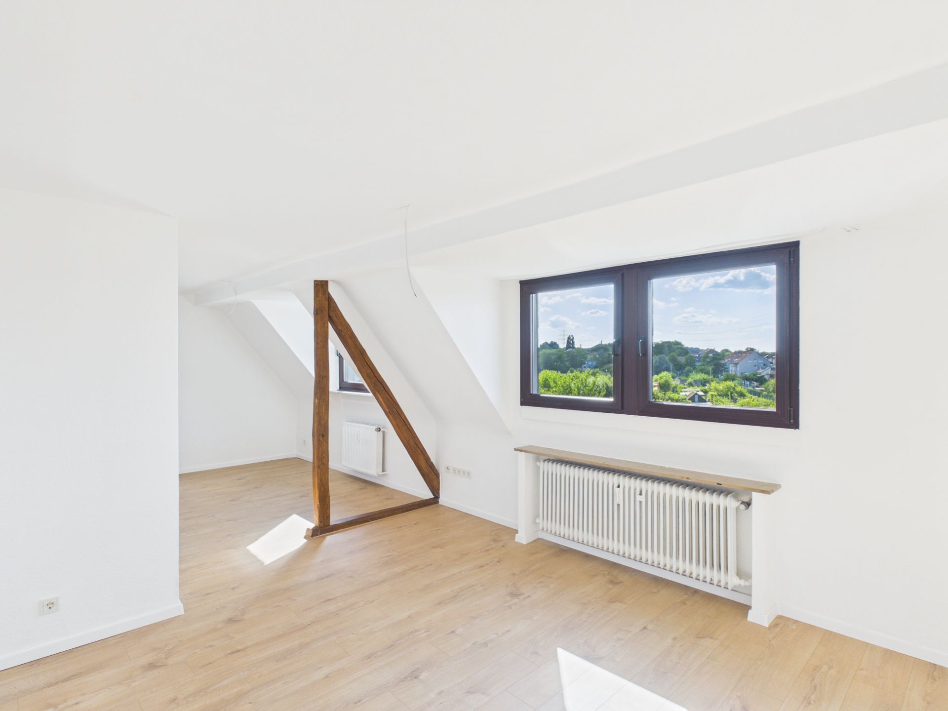 Helles Wohnzimmer mit Holzfußboden, Dachschräge, Holzbalken und großem Fenster mit Blick ins Grüne.