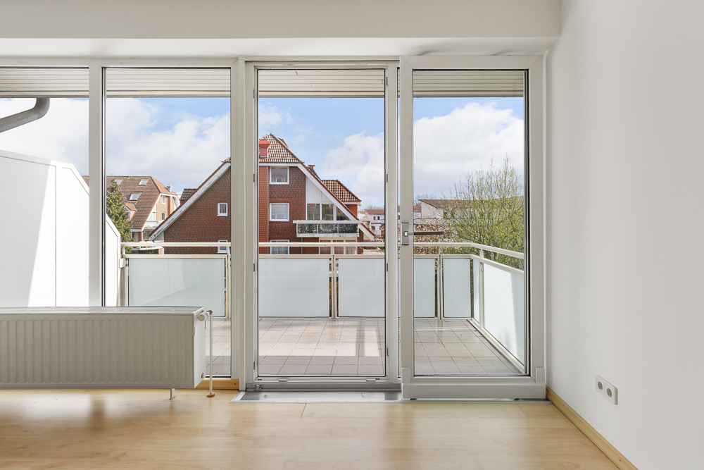 Blick aus einem hellen Zimmer auf einen Balkon mit Glasgeländer und Wohnhäuser im Hintergrund.