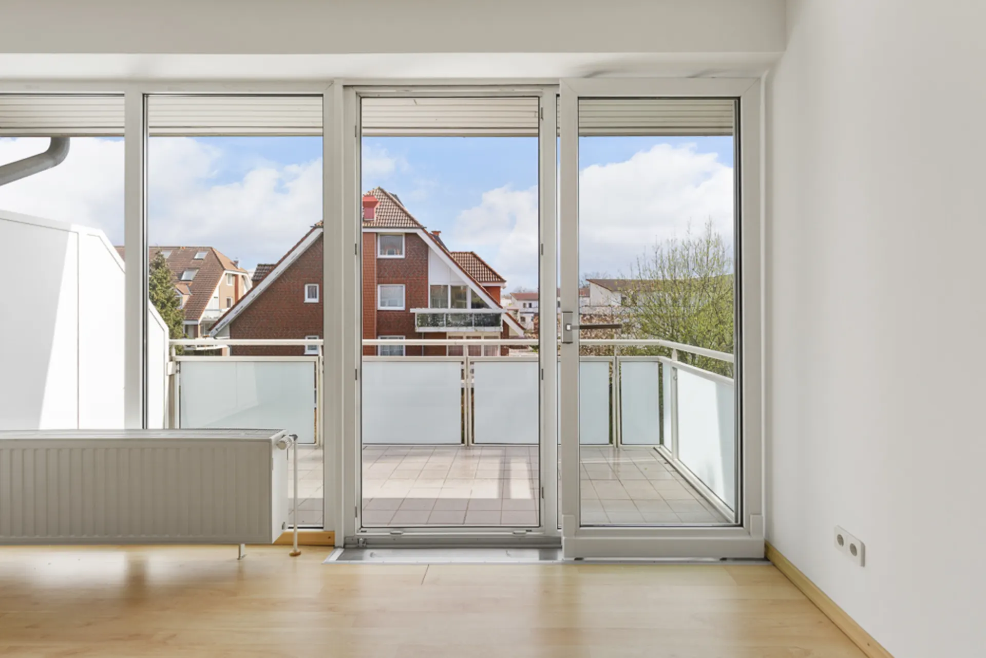 Blick aus einem hellen Zimmer auf einen Balkon mit Glasgeländer und Wohnhäuser im Hintergrund.