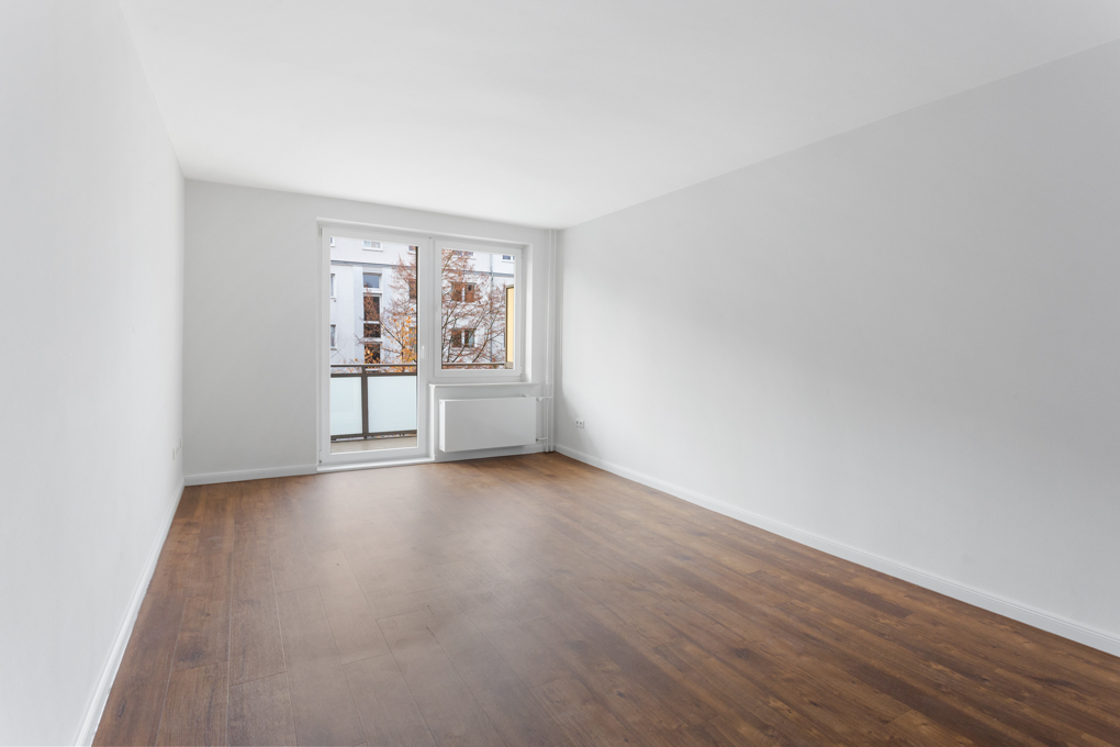 Leerer Raum mit weißem Wänden, braunem Boden und großen Fenstern zu einem Balkon.