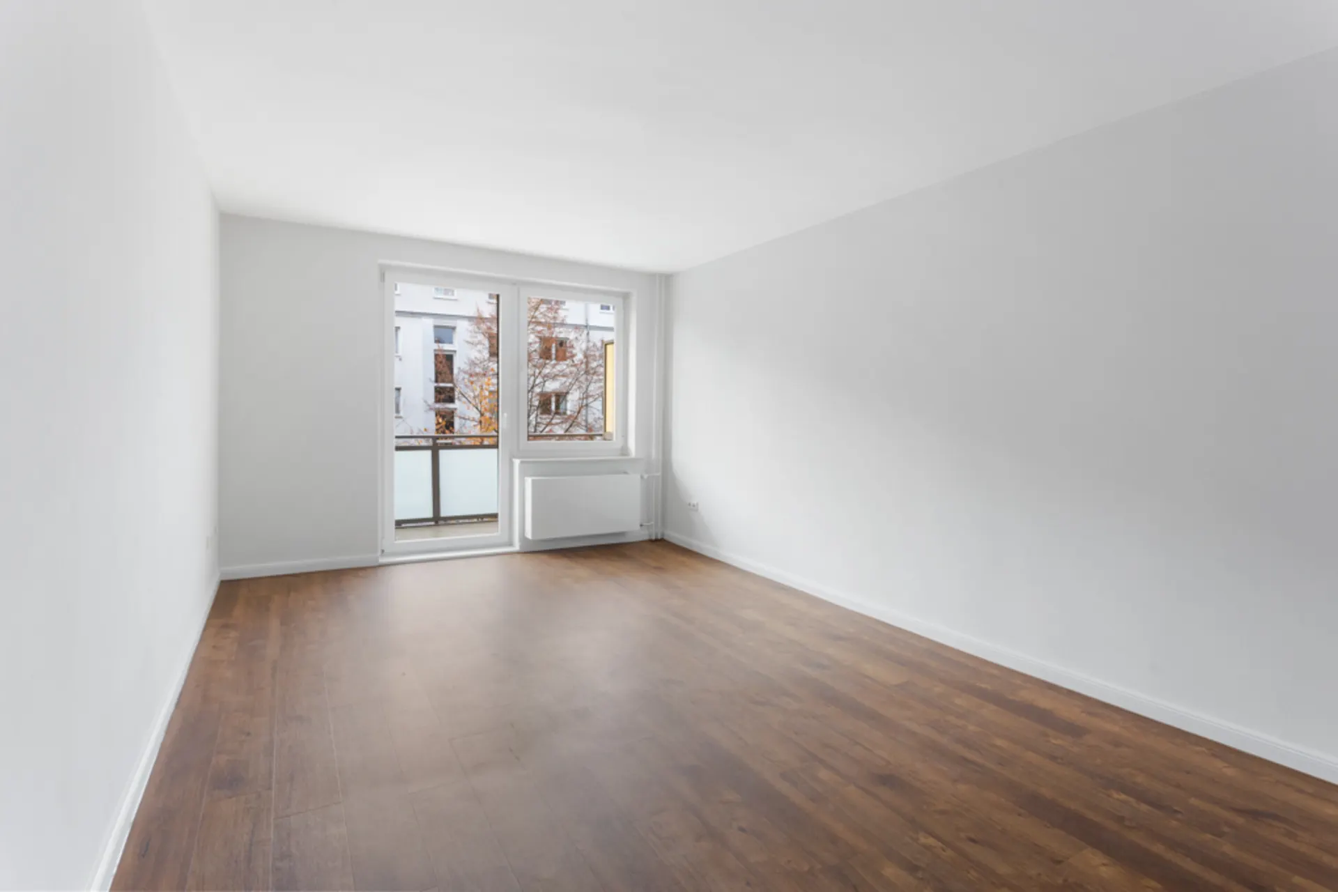 Leerer Raum mit weißem Wänden, braunem Boden und großen Fenstern zu einem Balkon.