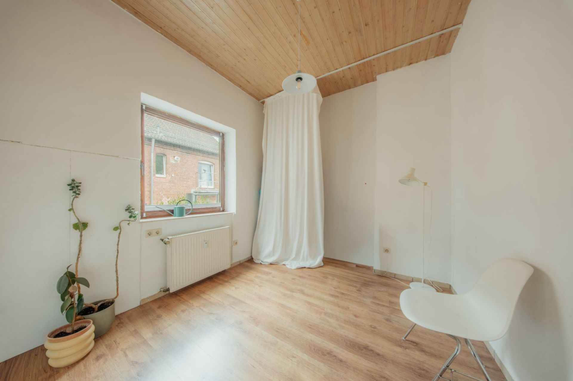 Helles Zimmer mit Holzdecke, Fenster, weißem Stuhl, Stehlampe, Pflanzen und Vorhang.