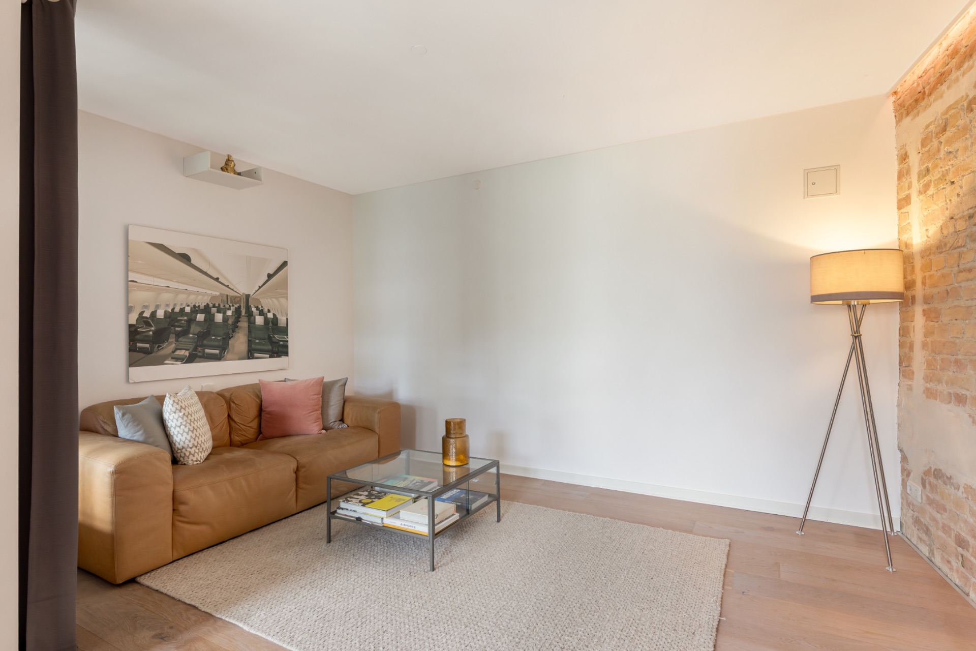 Wohnzimmer mit braunem Sofa, Glastisch, Stehlampe und Teppich auf Holzboden.