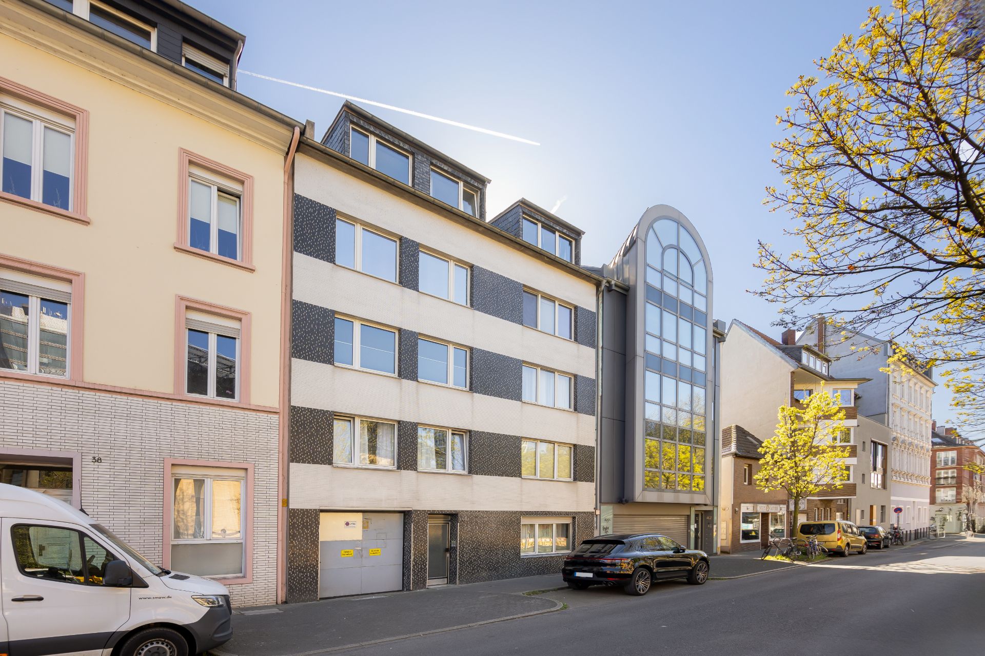 Mehrstöckiges Wohnhaus mit großen Fenstern an einer ruhigen Straße, Bäume und geparkte Autos sichtbar.