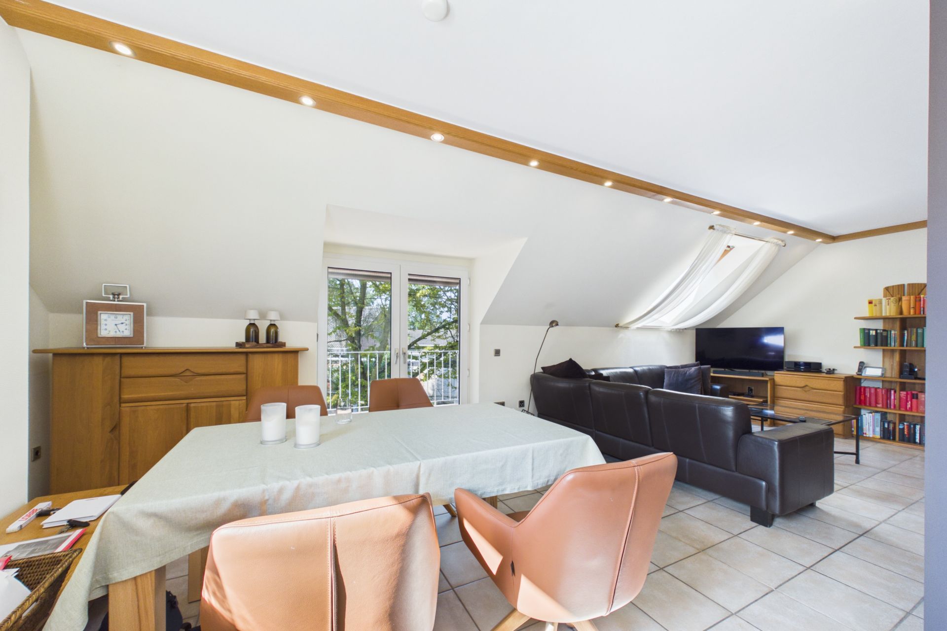Helles Wohnzimmer mit Esstisch, braunen Stühlen, Sofa, Fernseher und Bücherregalen.