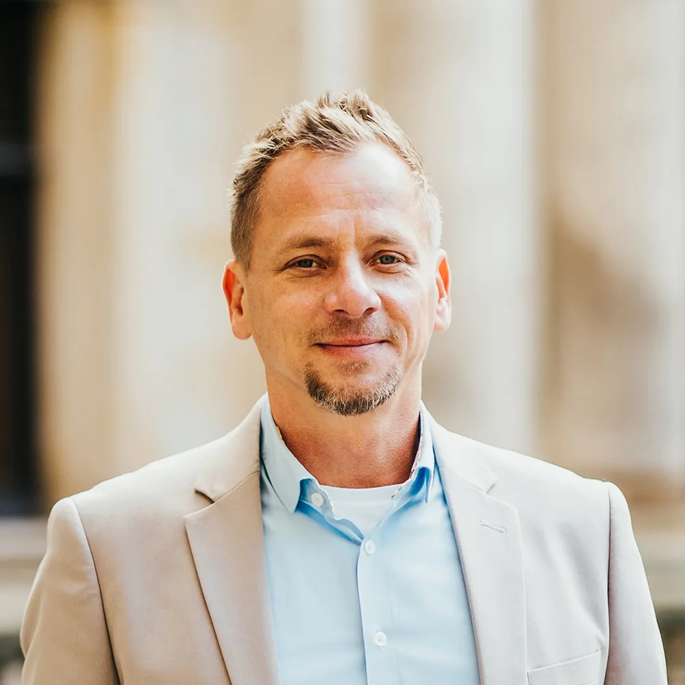 Ein Mann mit blondem Haar trägt ein beiges Sakko und ein hellblaues Hemd und steht vor unscharfem Hintergrund.