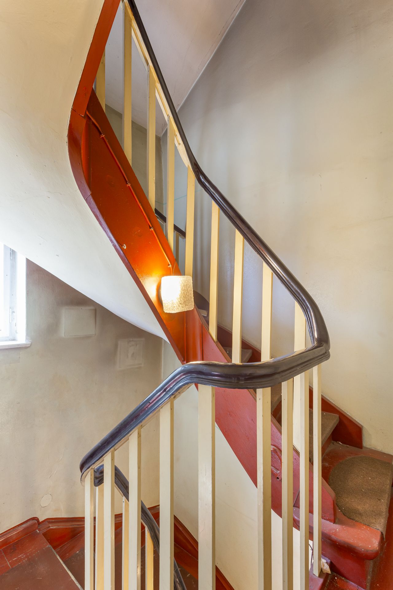 Rote Holztreppe mit cremefarbenem Geländer in einem Wohnhaus, beleuchtet von einer Wandlampe.