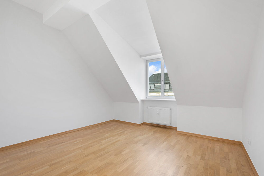 Leeres Zimmer mit weiß gestrichenen Wänden, Dachschrägen, Fenster und hellem Holzboden.