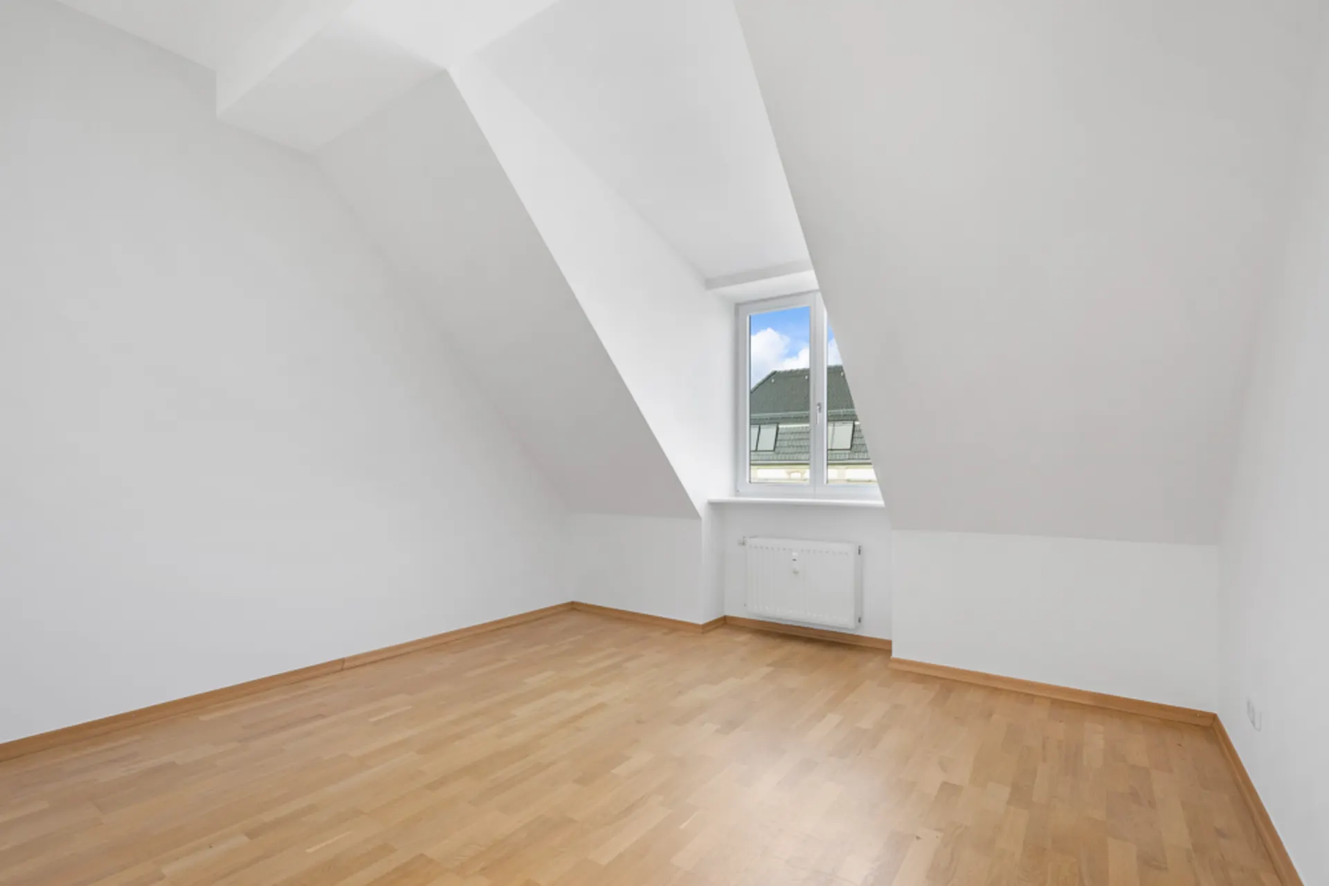 Leeres Zimmer mit weiß gestrichenen Wänden, Dachschrägen, Fenster und hellem Holzboden.