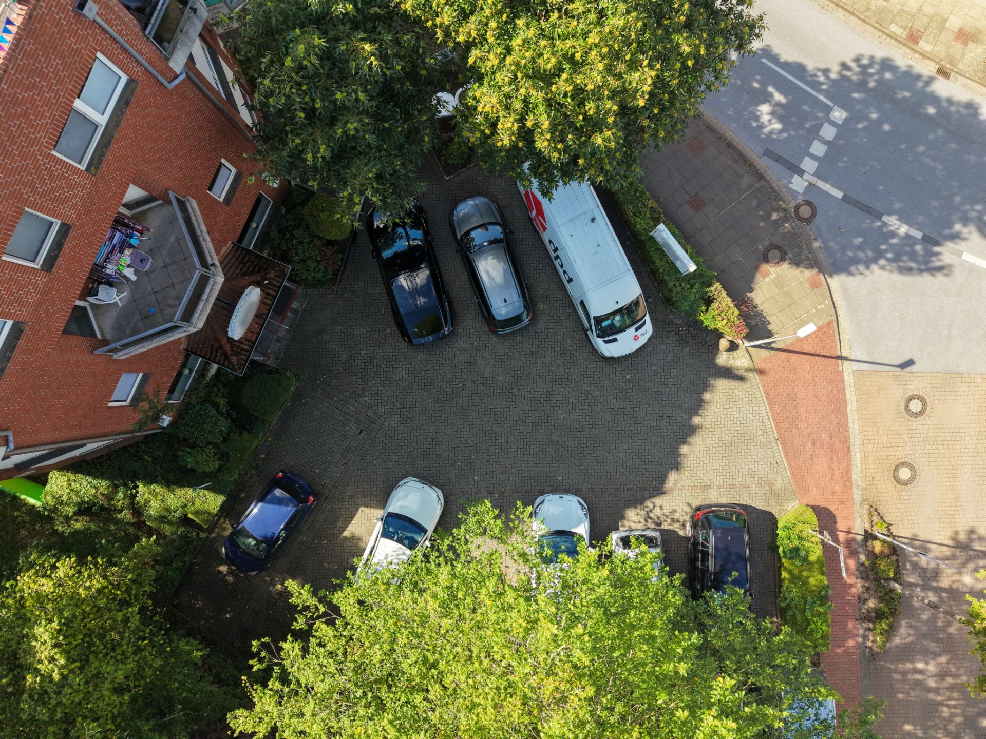 Luftaufnahme eines Parkplatzes mit mehreren Autos neben einem Wohngebäude und Bäumen.