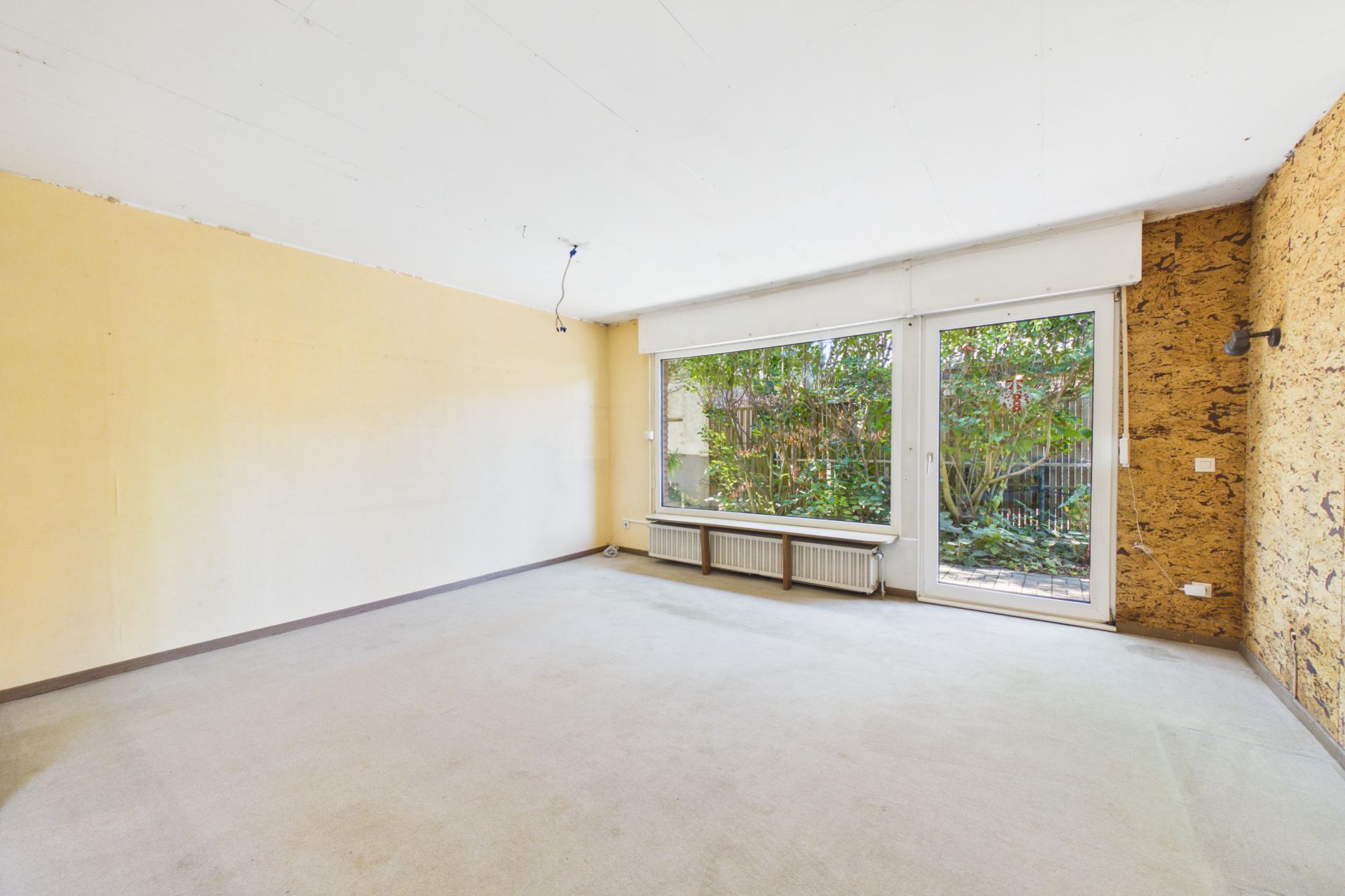 Leeres Zimmer mit großem Fenster und Tür zum Garten, helle Wände und Teppichboden.