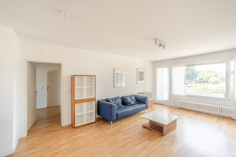 Helles Wohnzimmer mit Holzboden, blauem Sofa, Couchtisch und Regal, große Fenster mit Blick ins Grüne.